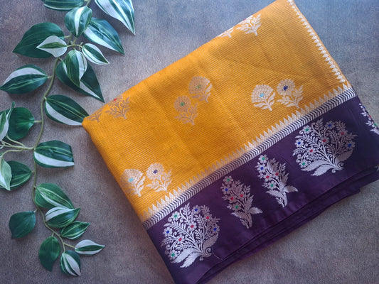 Purple and yellow saree with floral patterns on a textured surface with green leaves.