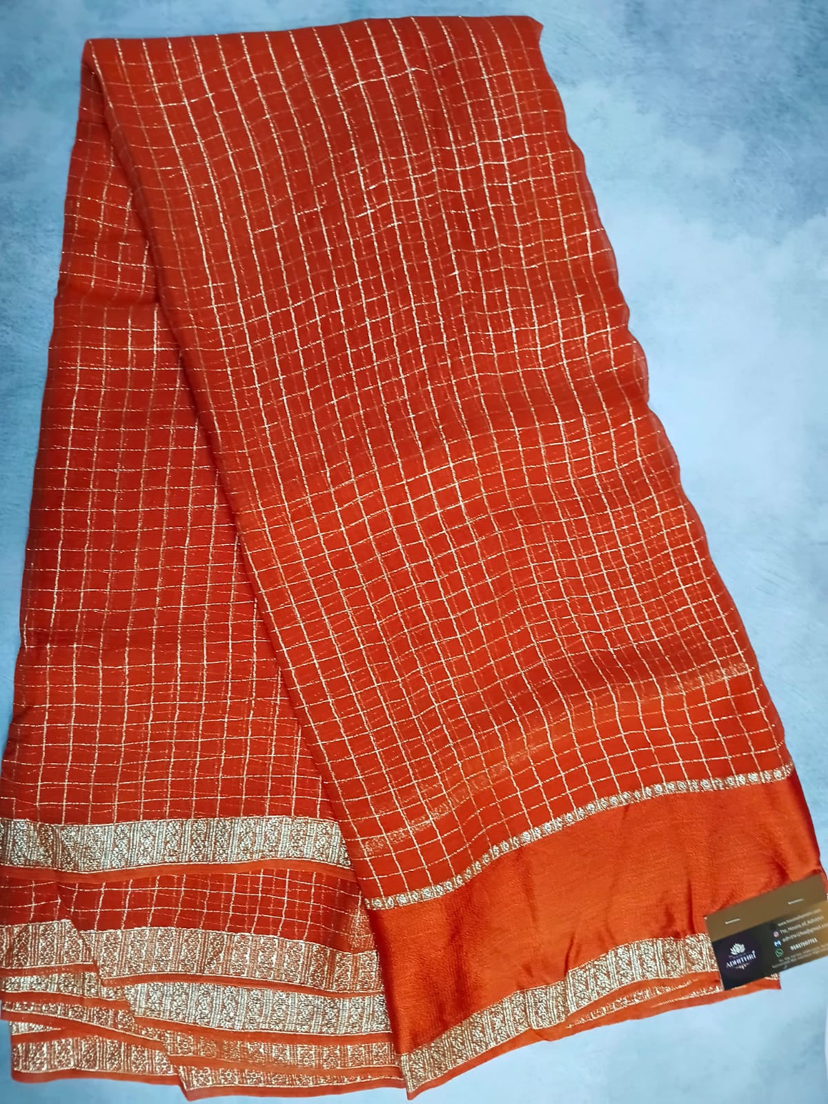 Orange saree with white checkered pattern and gold border on a light blue background
